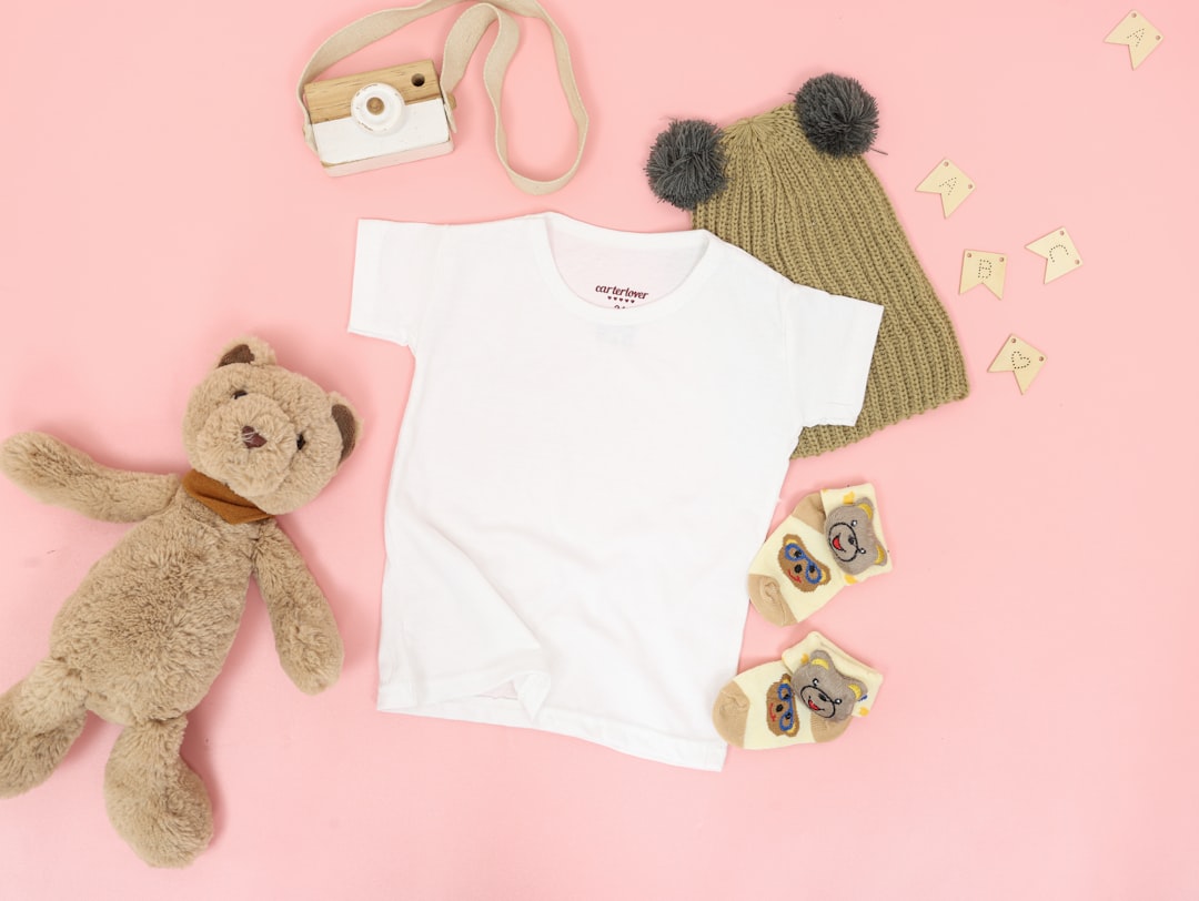 Blank white baby clothes lying on a pink surface next to a teddy bear and a cute bear hat.